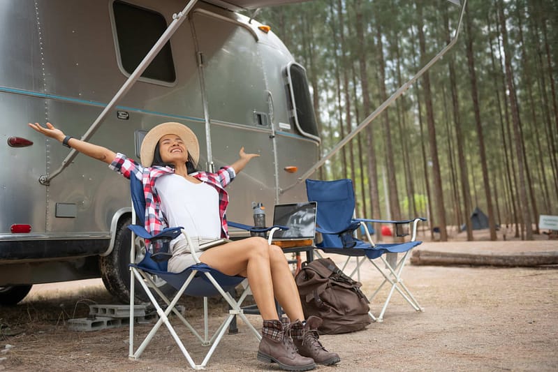 A woman in a hat sits stretching joyfully in a chair outside a camper van in an auto draft forested campground.