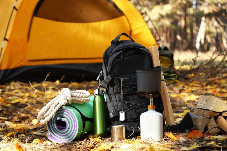 Camping gear, including a backpack, rolled-up sleeping mat, rope, thermos, cup, camp stove, and an axe, is meticulously arranged in front of a pitched yellow tent amongst fallen leaves in a forest setting. This scene feels like the perfect auto draft for your next adventure.