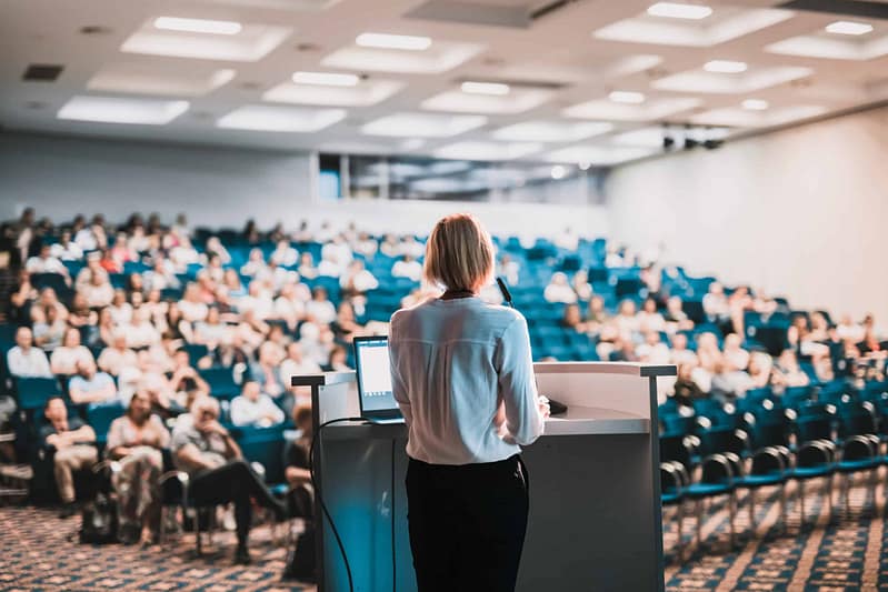 A woman presenting at a podium in a conference room filled with attendees from the camping industry.