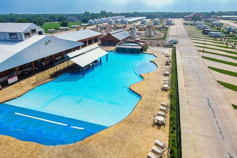 A large, outdoor swimming pool is adjacent to a pavilion with a covered area. Lounge chairs line one side of the pool, and buildings and a road are visible in the background, giving it an auto draft atmosphere.