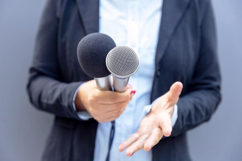 A woman is holding two microphones while providing public comment at an Oregon State Parks Dept meeting regarding increased out-of-state RV fees.