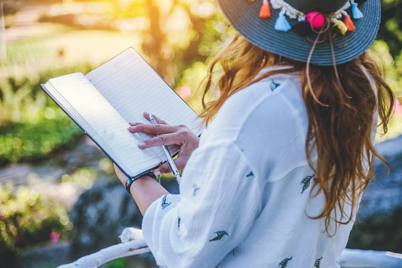 A woman wearing a hat is participating in the Writing Contest by Share Your Story as she writes in a notebook.
