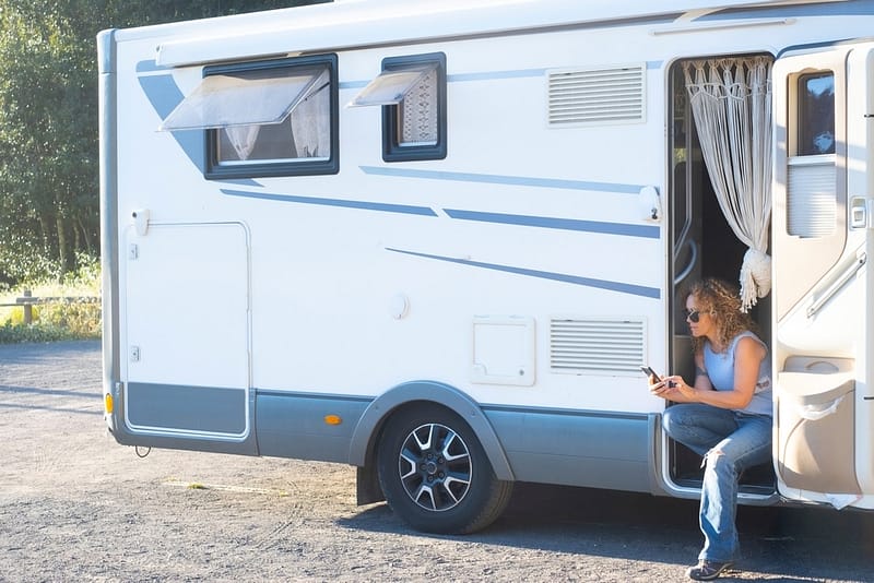 A woman sitting outside of an RV, engrossed in her phone while browsing through the KOA Report.