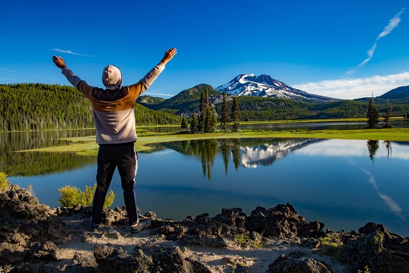 A man is standing on a rock in Oregon with his arms outstretched in front of a lake.