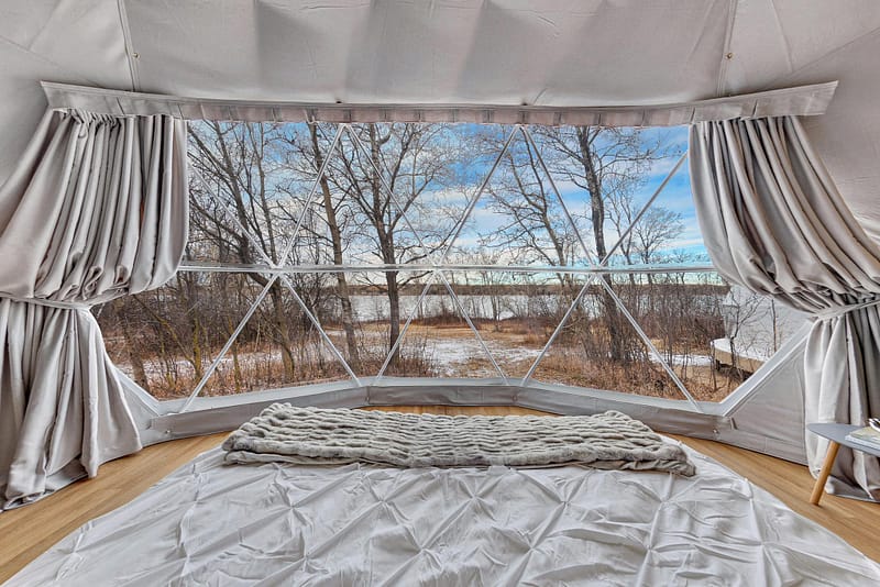 An interior view of a tent at a glamping resort with a large window showing a wintry outdoor scene with bare trees and light snow on the ground. A bed with white linens and a knitted blanket lies near the window, offering cozy comfort while you gaze at Blackstrap Lake.