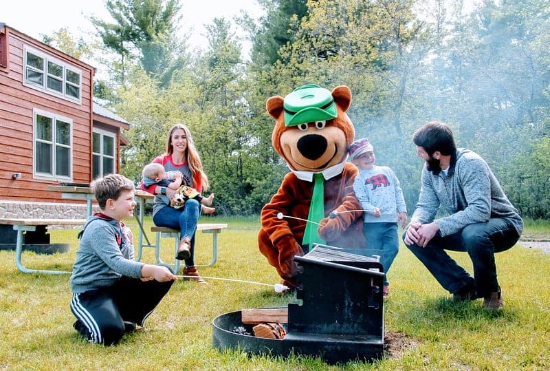 A family is sitting around a campfire at a Jellystone Park, with a bear mascot.