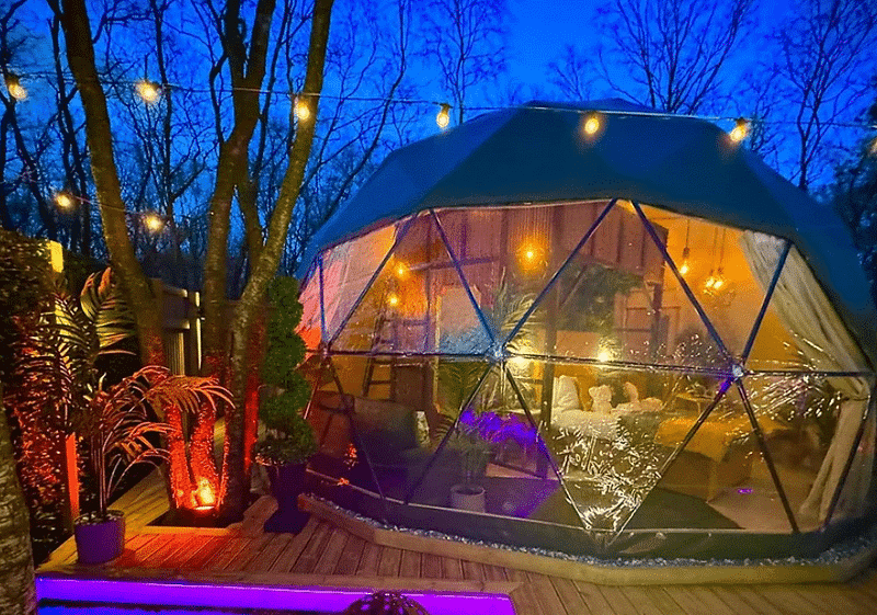 A geodesic dome illuminated by string lights at dusk, featuring a cozy interior and surrounded by a wooden deck and lush plants.
