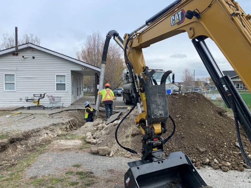 A construction worker supervises an excavator digging near a house, with piles of dirt and auto draft equipment scattered around.