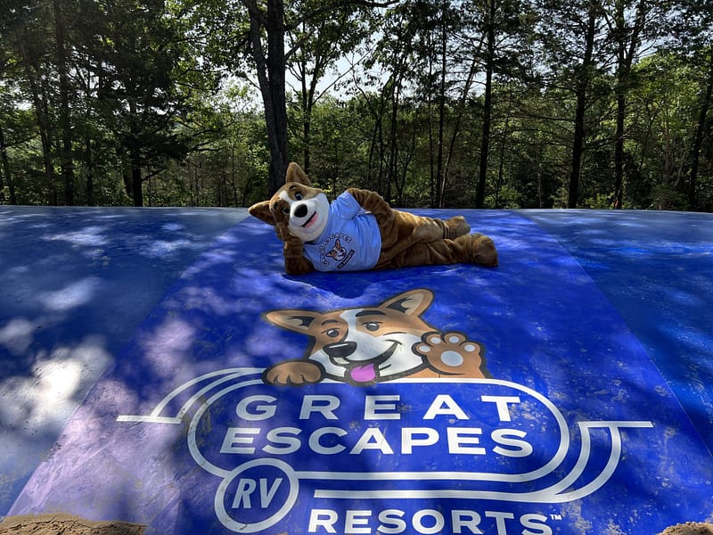 A costumed mascot, resembling a dog, lies on an inflated blue surface adorned with the "Great Escapes RV Resorts" logo, as trees sway gently in the background. Auto Draft
