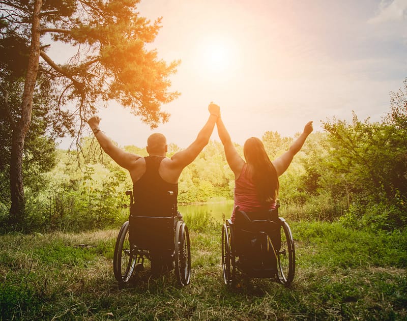 Two people in wheelchairs raising their hands in the Great Outdoors.