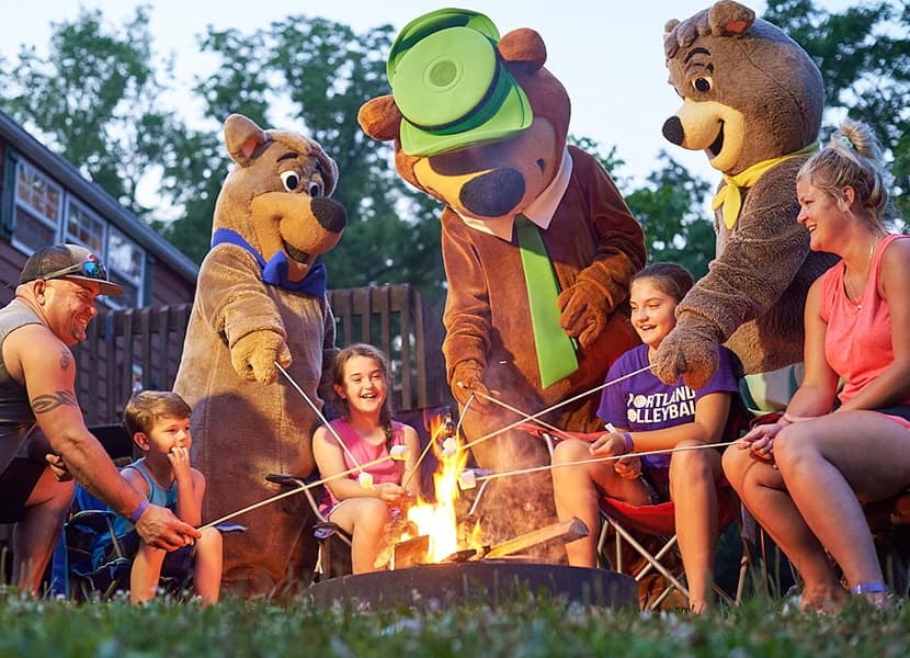 A family enjoys a campfire with two people in mascot costumes of bear characters, created from an Auto Draft.