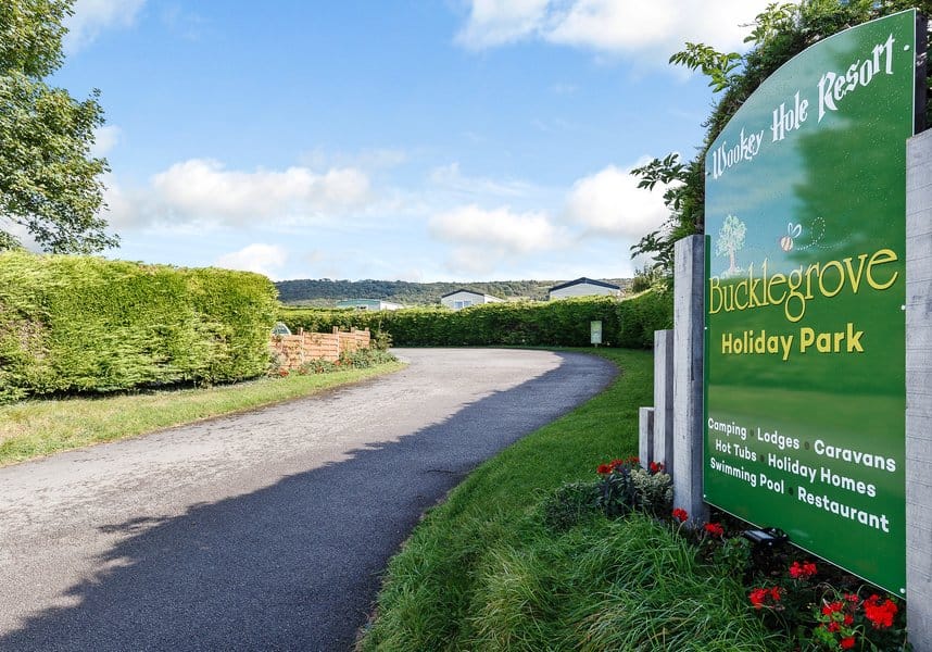 Entrance to Bucklegrove Holiday Park with signs and an auto-drafted paved road leading into the resort, surrounded by lush greenery and hills in the background.
