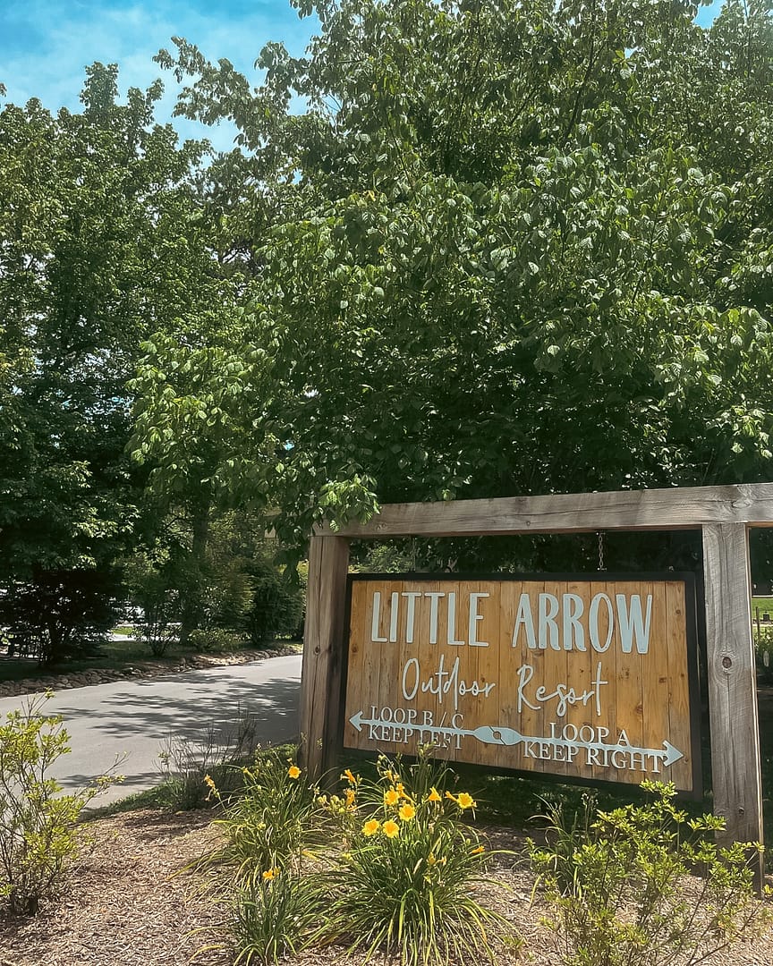 A sign for little arrow in front of bushes and trees at Little Arrow Outdoor Resort.