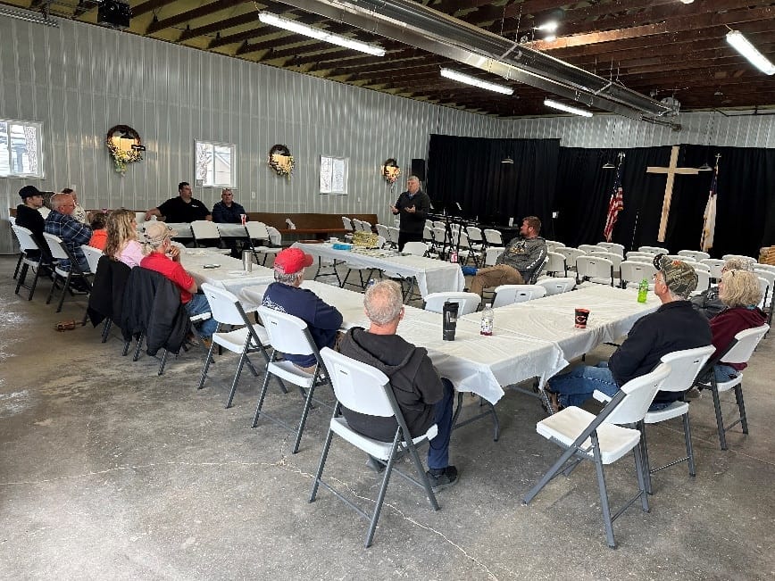 A group of people seated at tables in a large hall listening to a speaker at the front near a cross and American flag during the Illinois Campground Association's Statewide Spring Meetings.