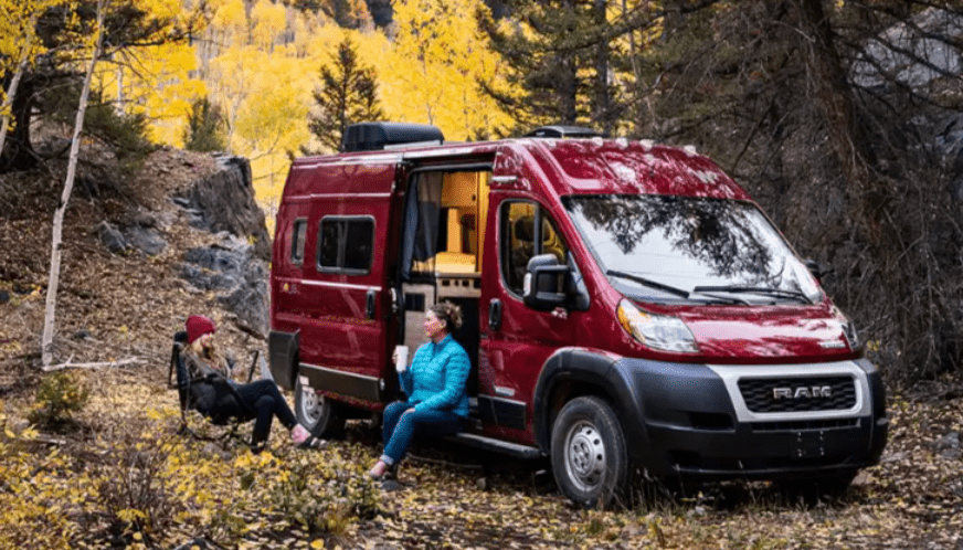A woman sits next to a red camper van, the Solis, in the woods.