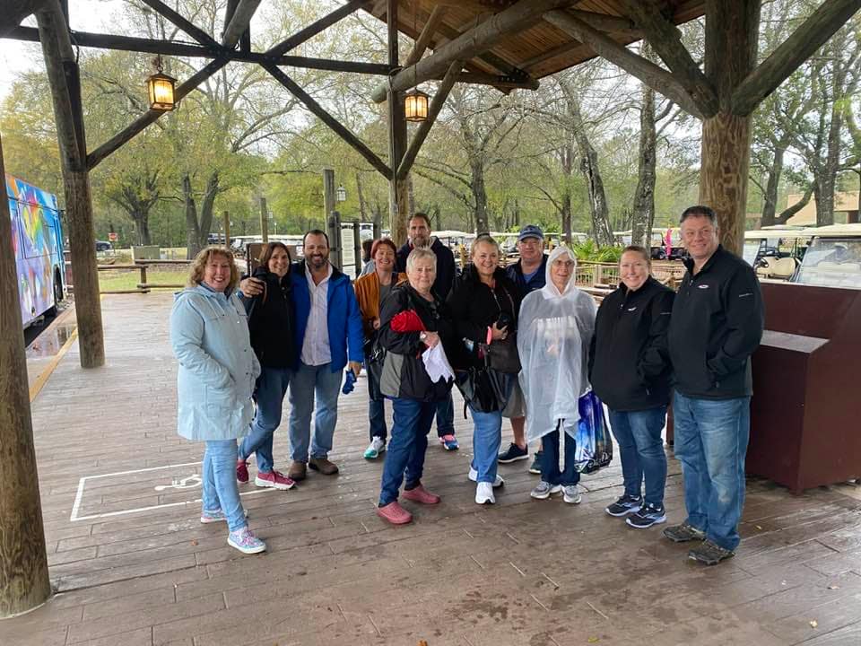 A group of adults standing under a wooden pavilion in a park, smiling at the camera on a cloudy day.