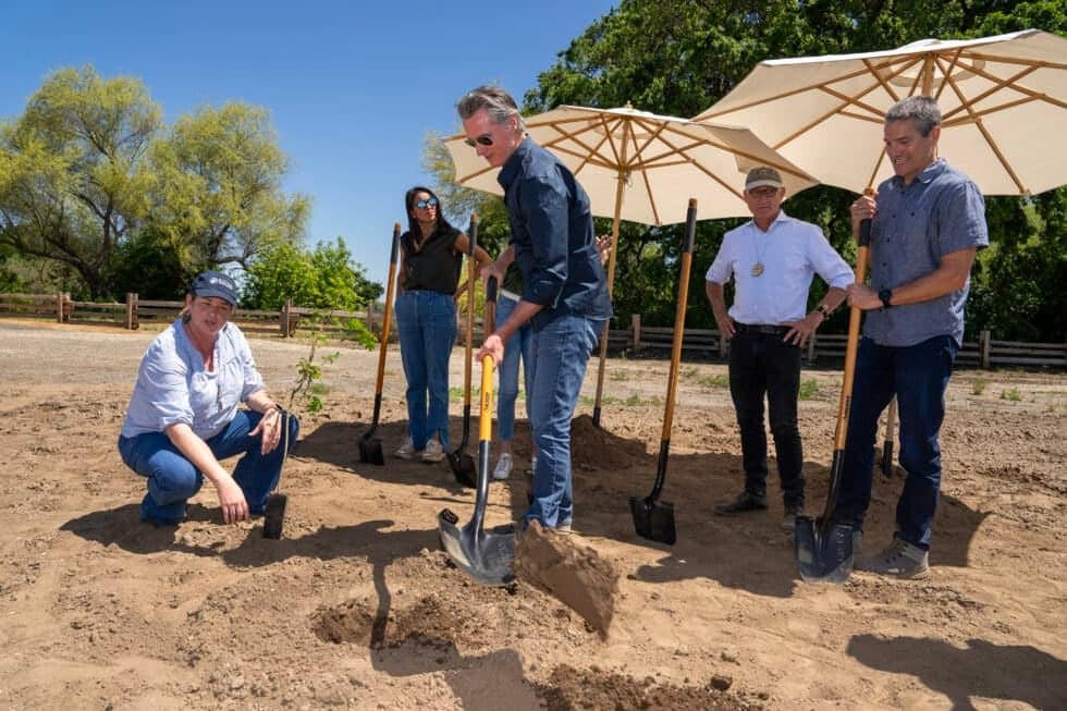 A group of five people, including one kneeling, engage in a tree-planting activity outdoors under parasols on a sunny day at a new California state park.