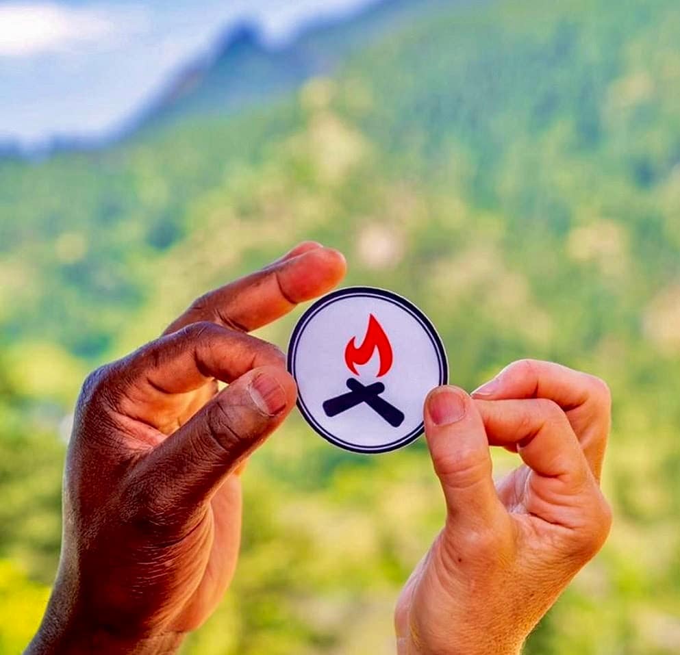 A person holding up a sticker with a campfire on it promoting the Black Folks Camp Too initiative.