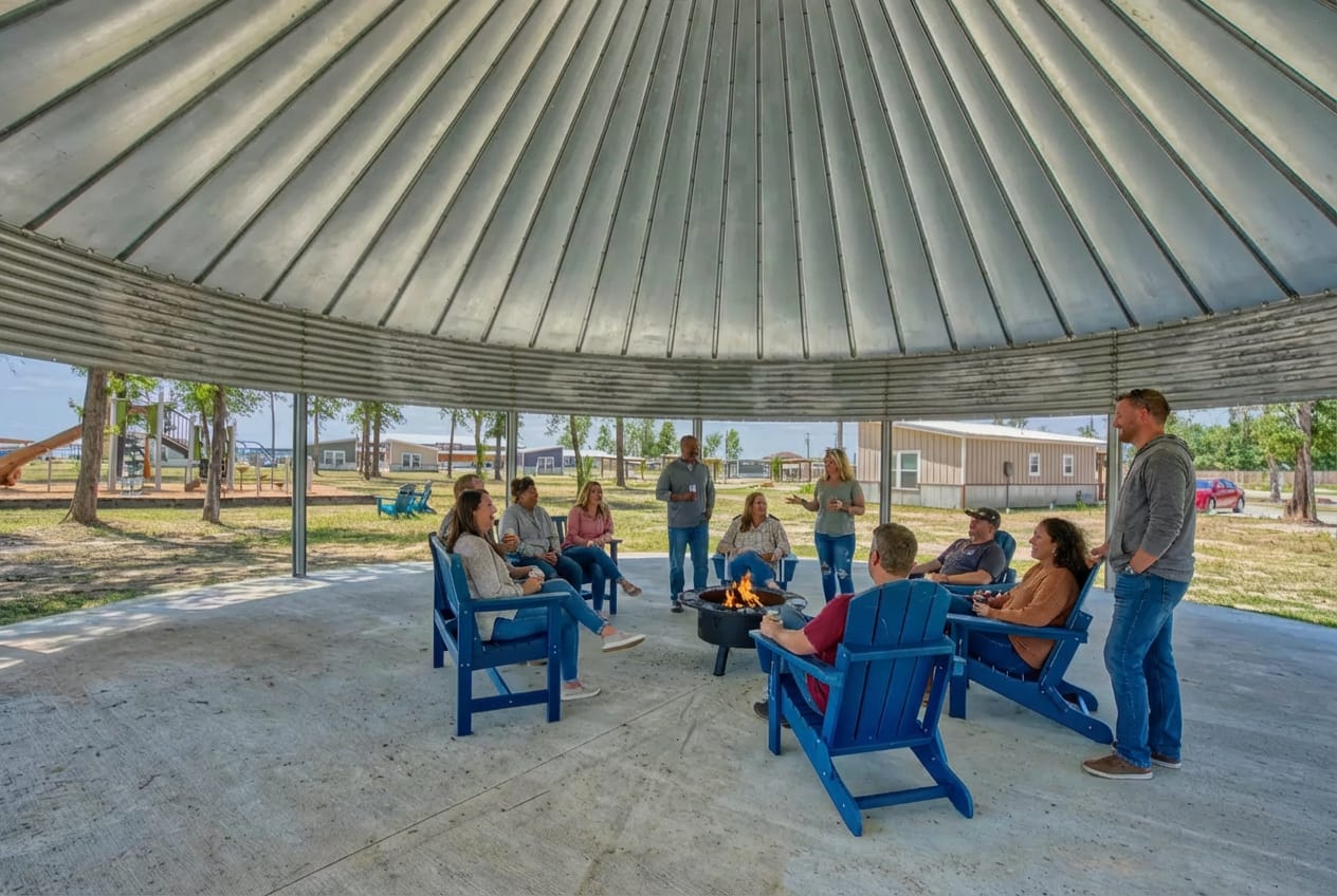 A group of people sit in blue chairs around a fire pit under a large open-sided metal-roofed pavilion on a concrete floor.