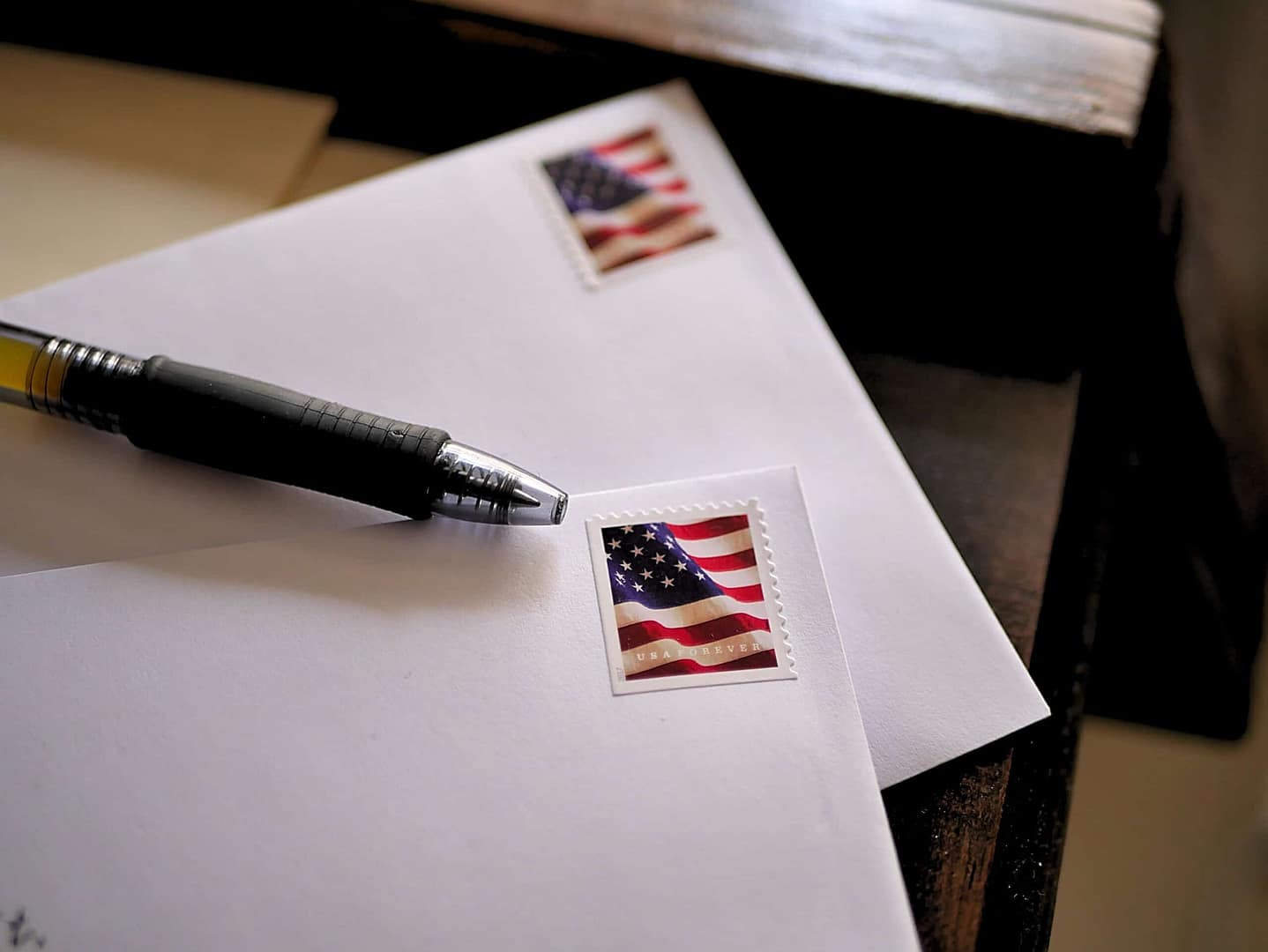 A close-up of two white envelopes on a dark wooden surface, each with a U.S. flag stamp. A black pen rests on one envelope, partially covering the stamp.