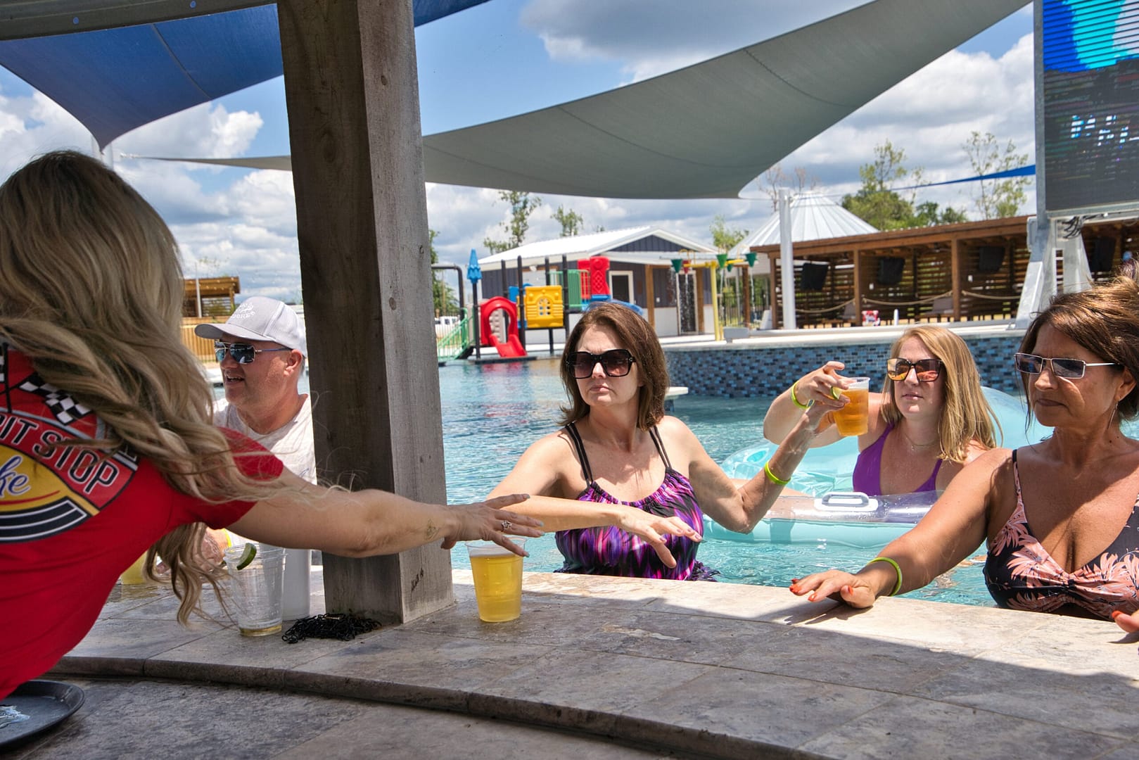 A bartender hands drinks to three women and a man seated at a swim-up bar in a pool on a sunny day.