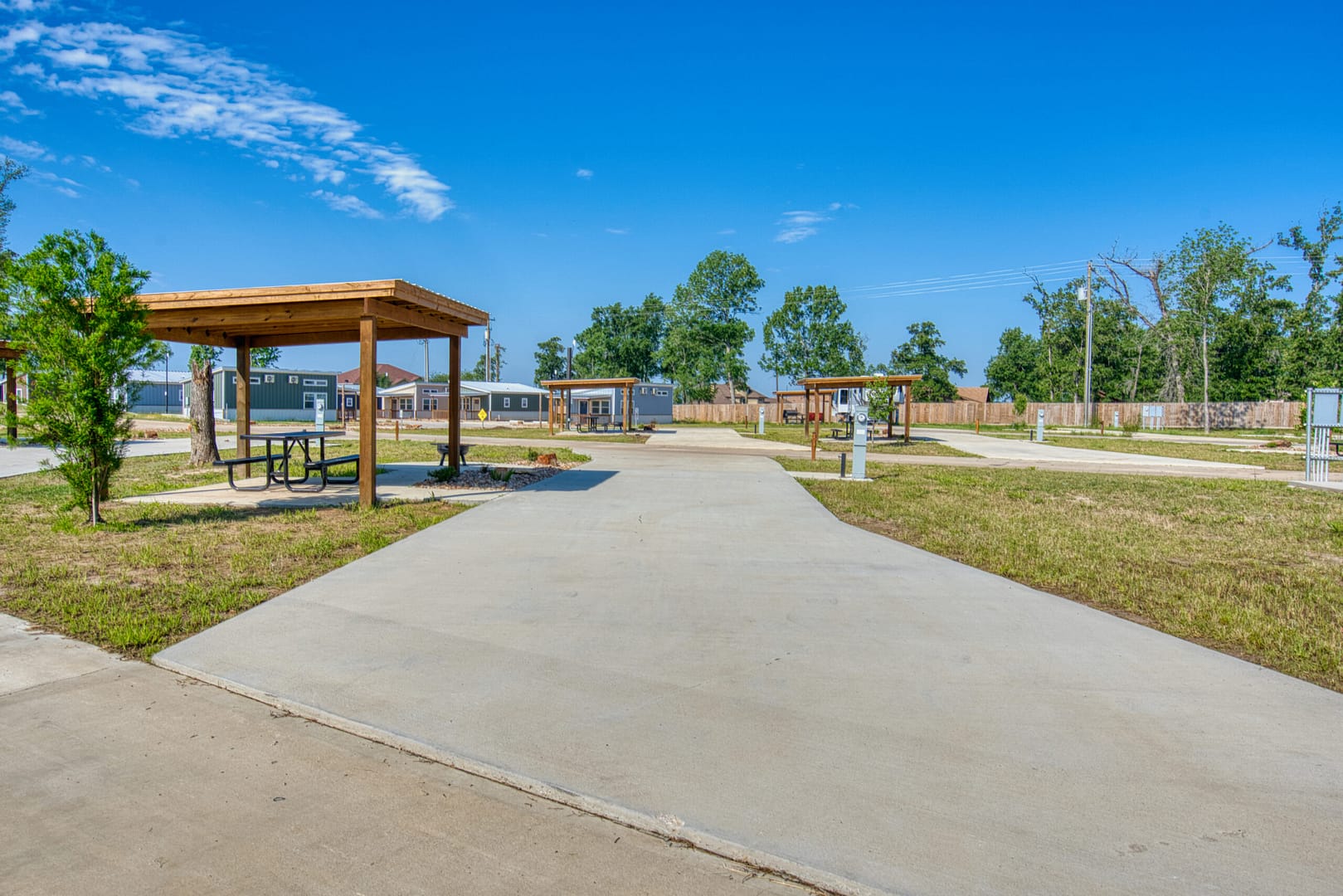Concrete RV parking pads with wooden picnic shelters and grassy areas under a clear blue sky at a campground.