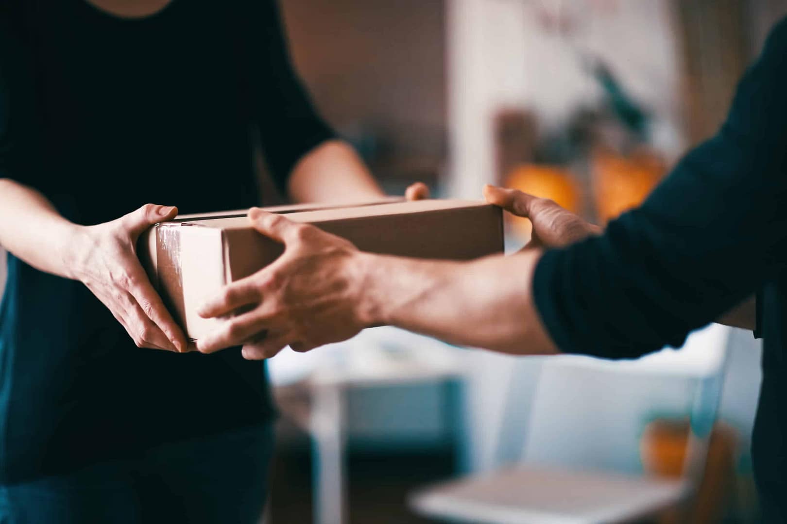 Two people exchanging a cardboard box indoors. One person is handing the box over while the other is receiving it. The background is blurred, with indistinct furniture and a warm color scheme.