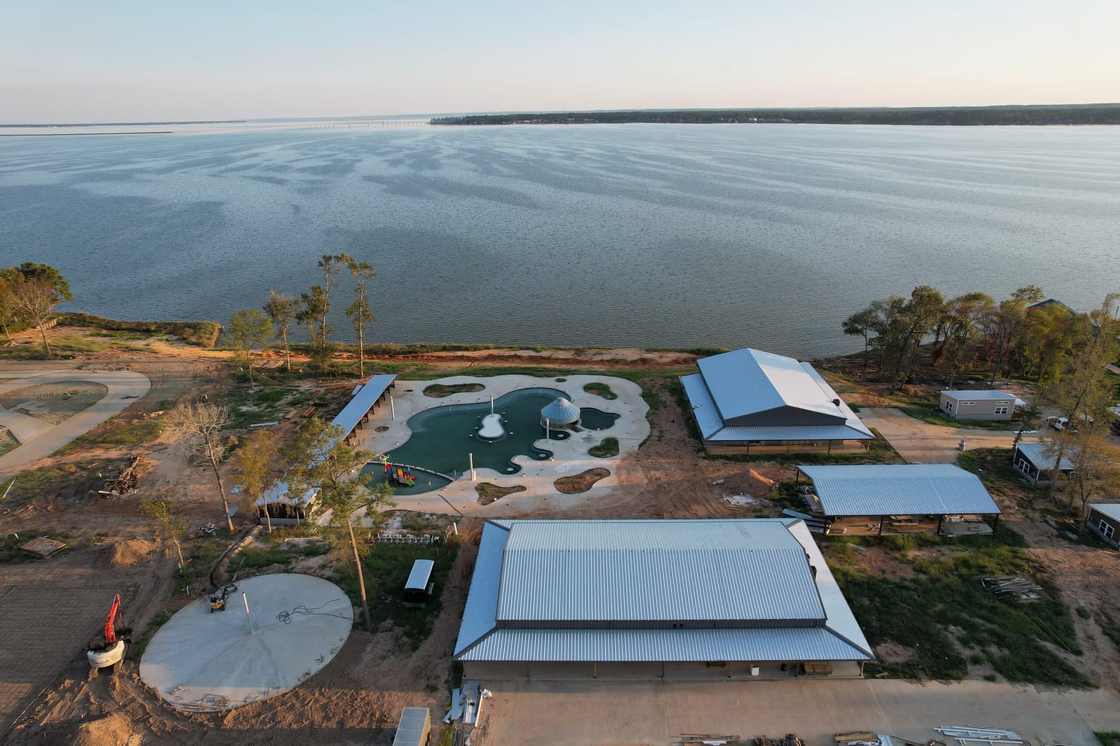 Aerial view of a lakeside area under development. Several buildings with metal roofs are visible, along with a circular structure and a pool surrounded by sand. The lake stretches across the upper half of the image under a clear sky.