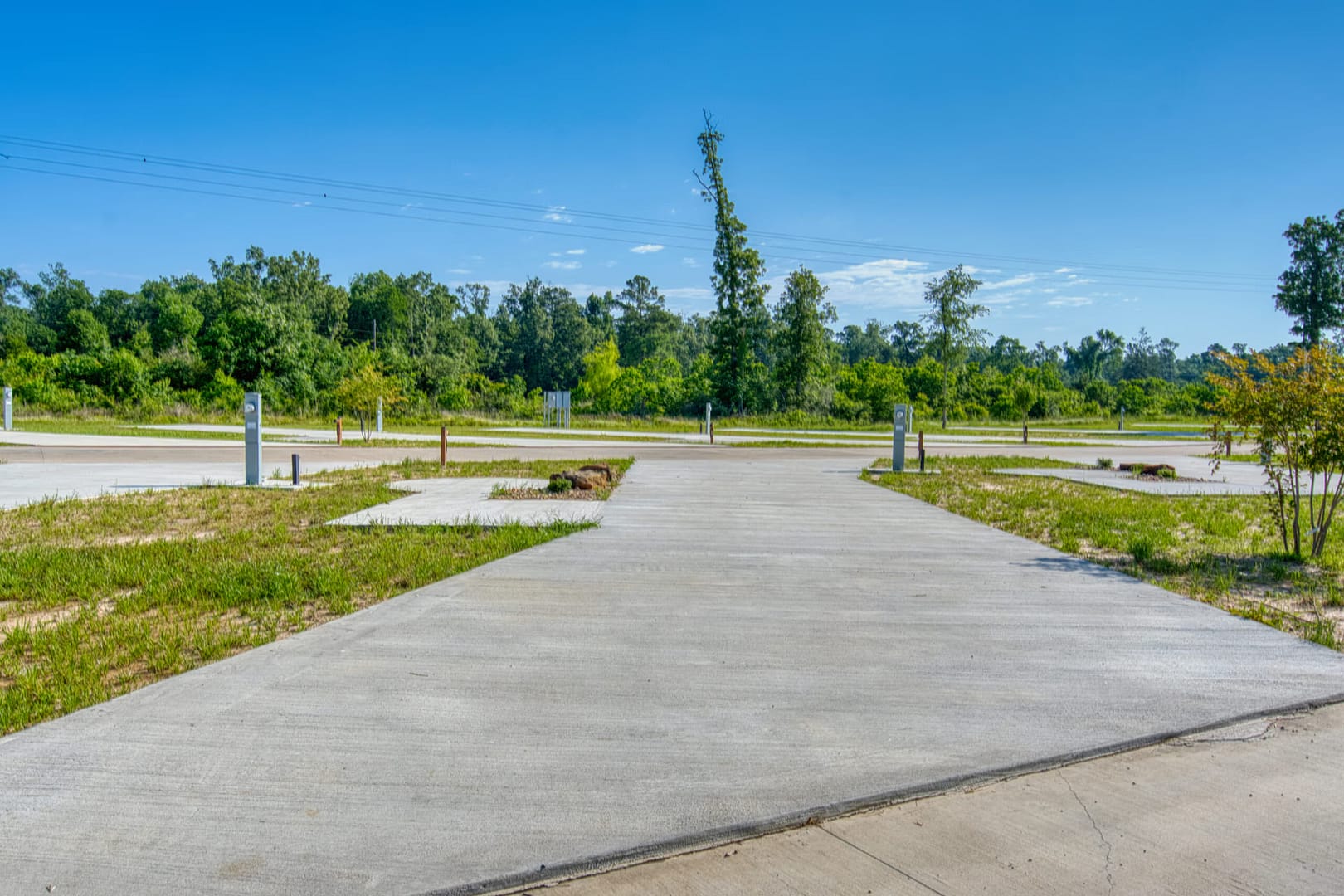 A wide, empty concrete lot with several utility hookups, surrounded by grass and trees under a clear blue sky. The area appears to be part of an RV park or campground.