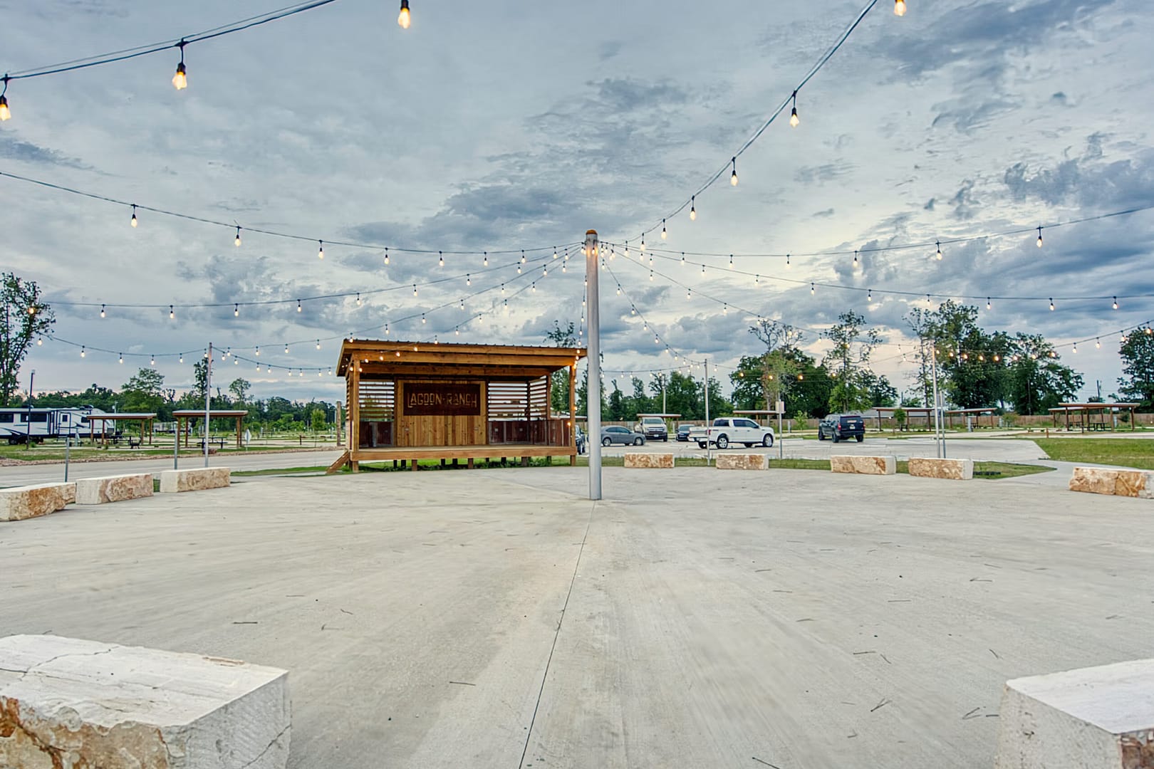 An outdoor pavilion with string lights overhead, surrounded by open concrete space and a few parked cars in the background, set against a cloudy sky and trees.