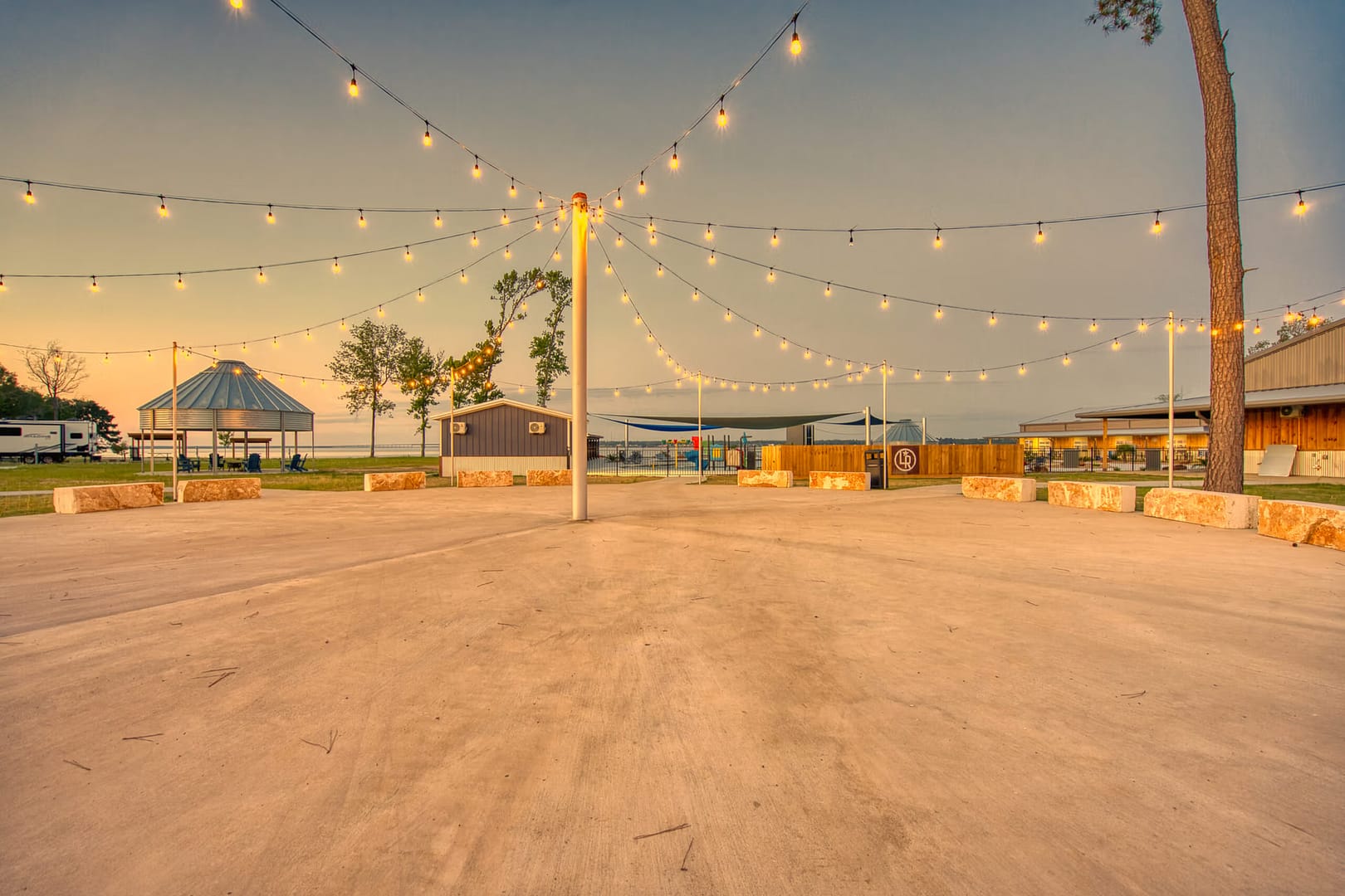 An outdoor venue with string lights overhead, stone benches arranged in a circle, and buildings and trees in the background at sunset.