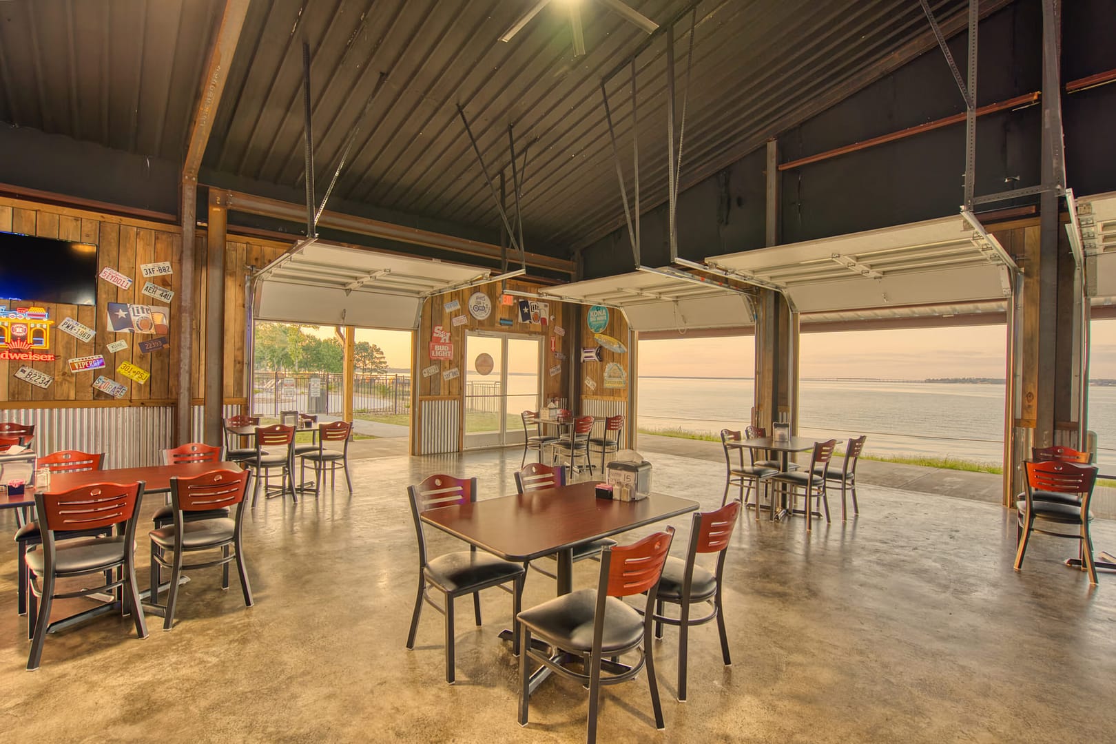 Open-air restaurant with red and black chairs, wooden walls, and large garage doors raised to reveal a waterfront view at sunset. Tables are spaced apart on a polished concrete floor.