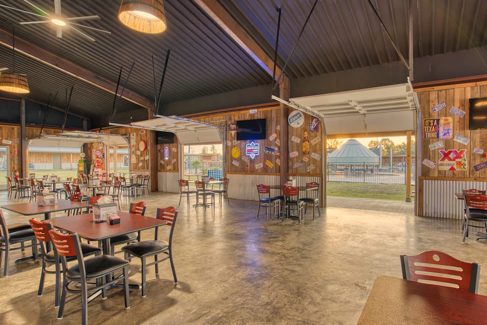 Open-air restaurant with wooden walls, large garage doors, various signs, and empty tables and chairs; outside view includes a pool area and trees at sunset.