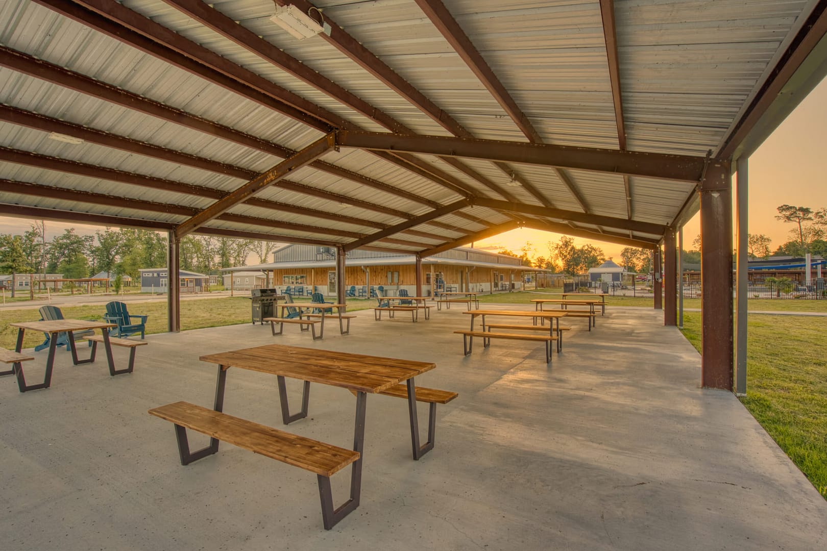 Open-air pavilion with a metal roof, several wooden picnic tables, and chairs on a concrete floor, with sunset light and grassy surroundings in the background.