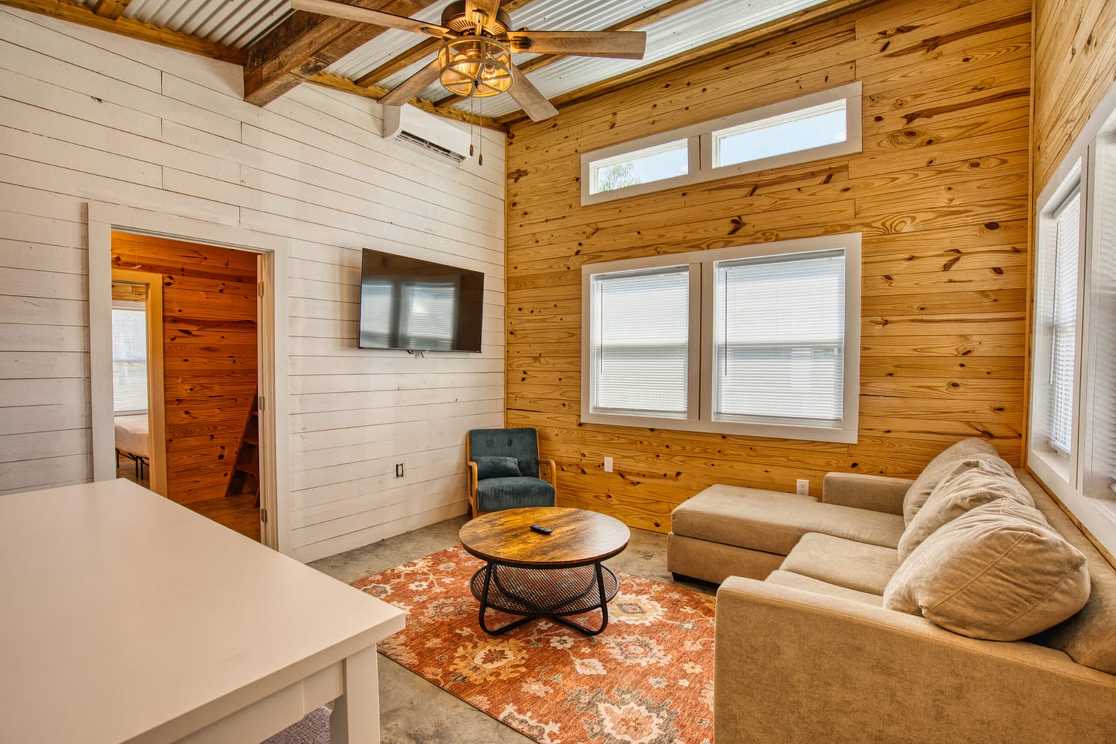A small living room with wood-paneled walls, a beige sectional sofa, round coffee table, wall-mounted TV, armchair, and an orange patterned rug under a ceiling with exposed beams.