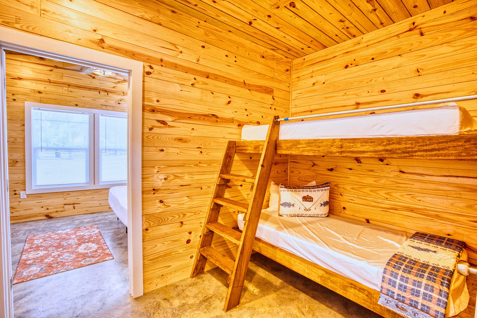 A wooden room with a bunk bed, ladder, fish-themed pillow, and a blue blanket; open doorway leads to a sunlit room with windows and an orange rug.