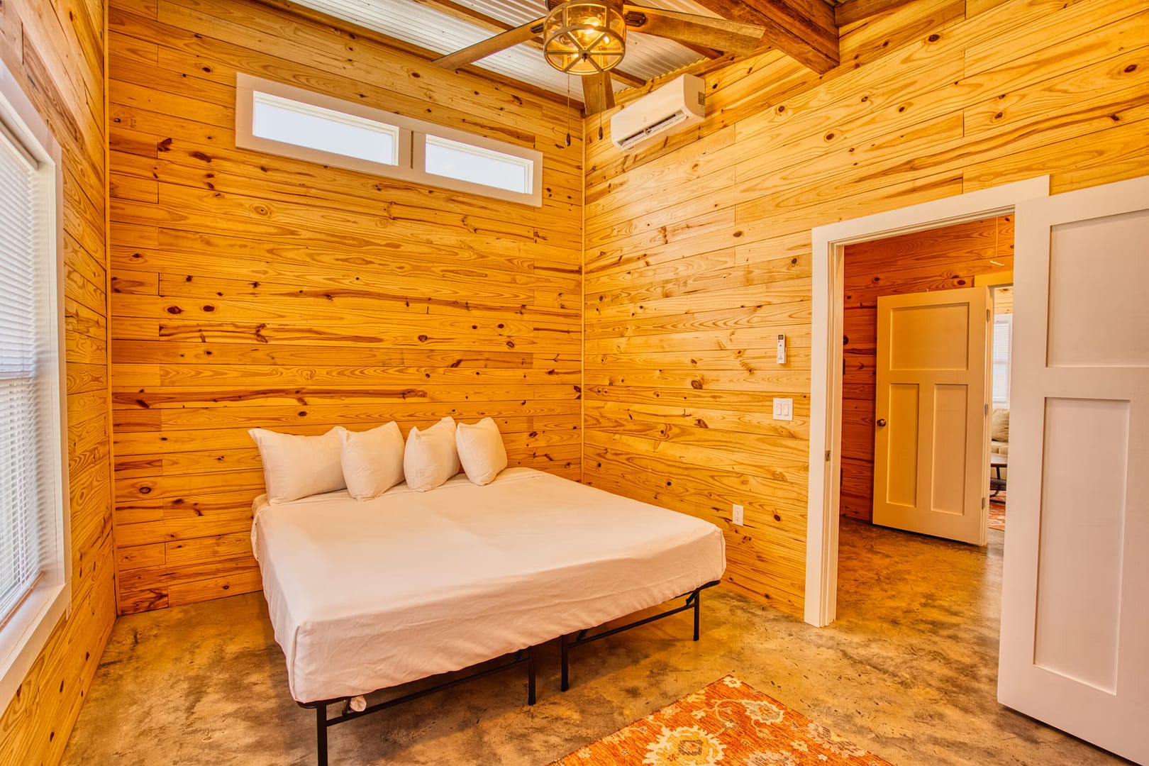 Bedroom with wood-paneled walls, a white bed with four pillows, concrete floor, ceiling beams, and a small orange rug near the bed.