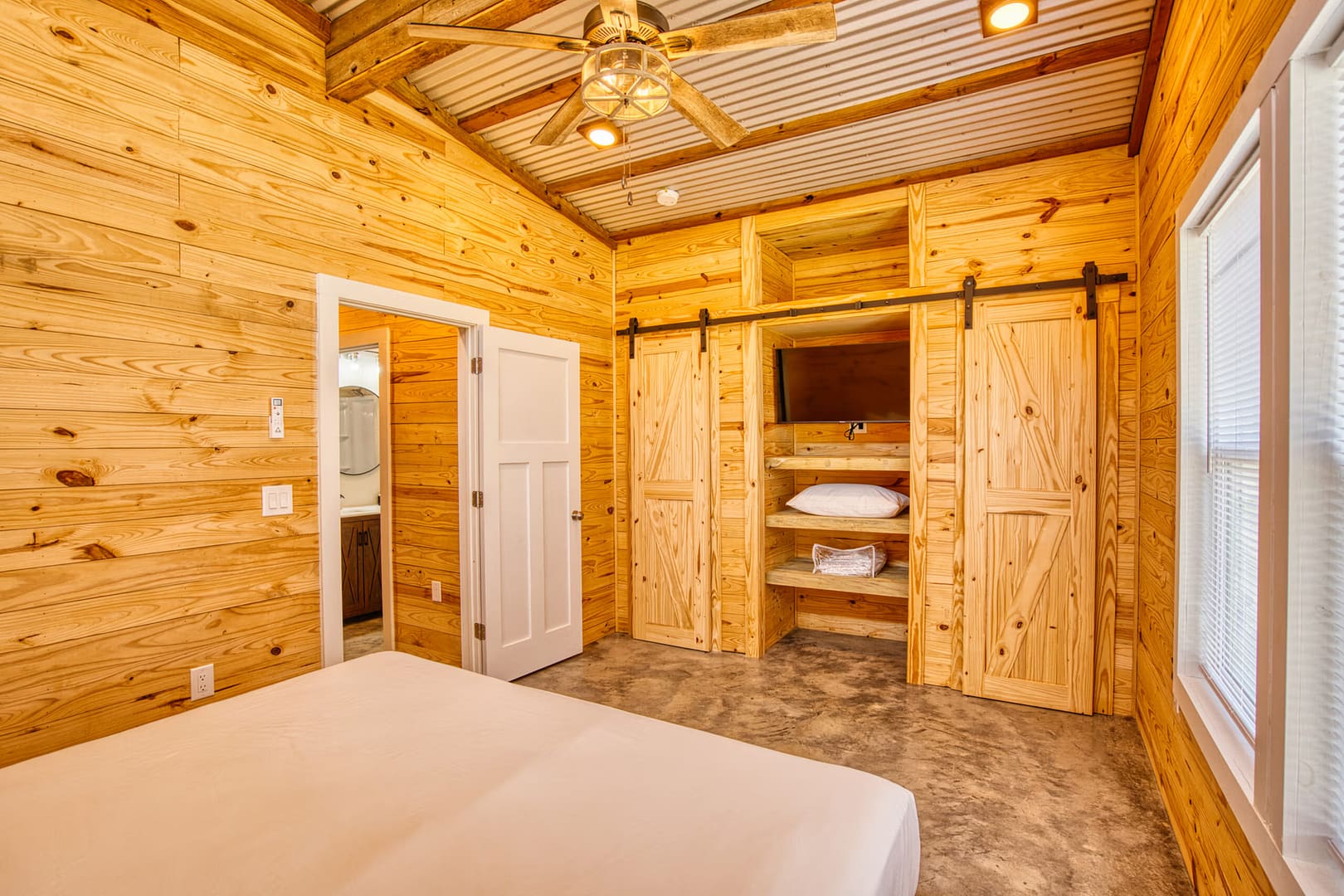 Bedroom with light wood paneling, a ceiling fan, a bed, an open closet with shelves, pillows, towels, and a wall-mounted TV. A door leads to a bathroom. Natural light from the window.