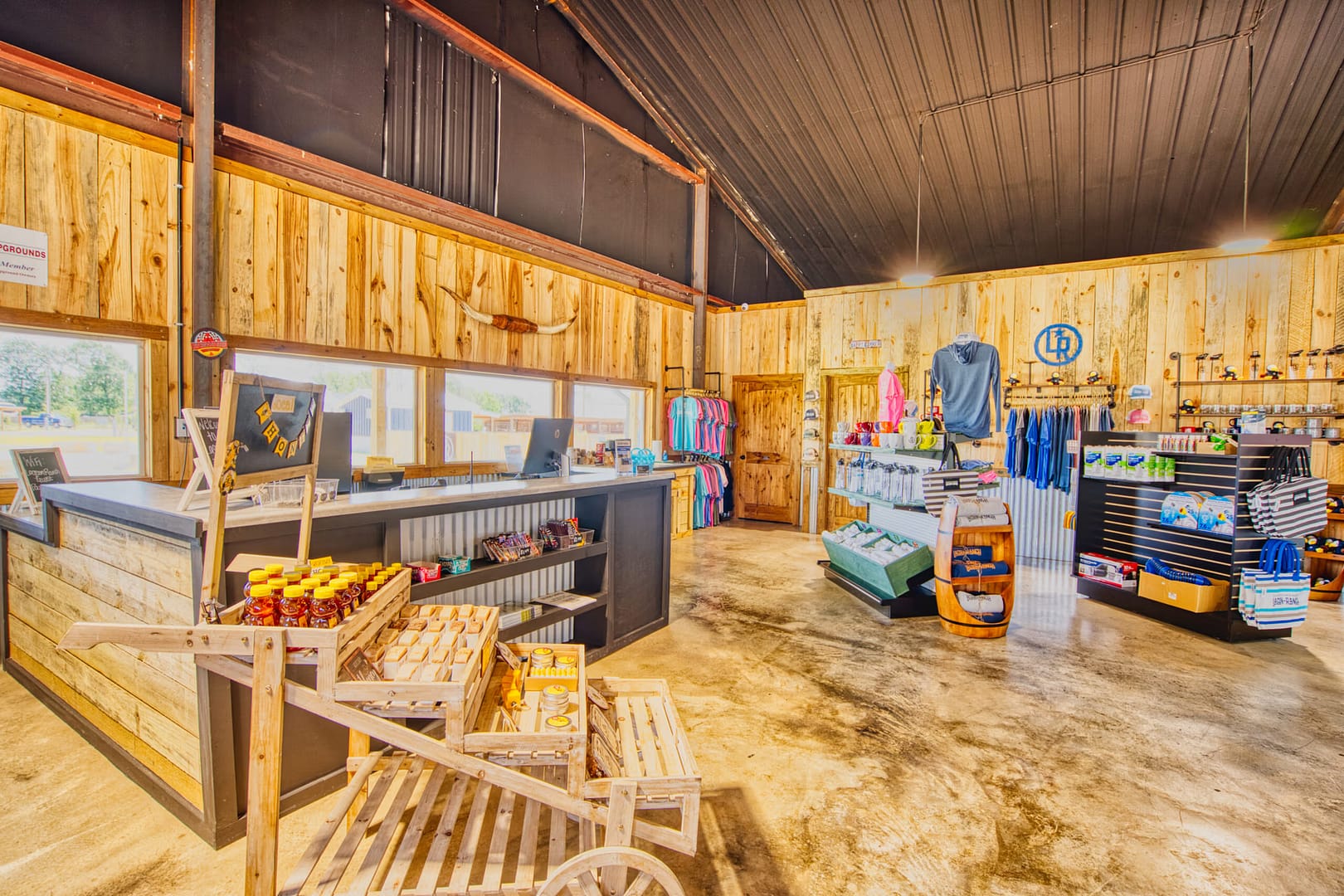A farm store interior with wooden walls, a counter, shelves displaying shirts, hats, honey, jam, and other products under bright lighting.