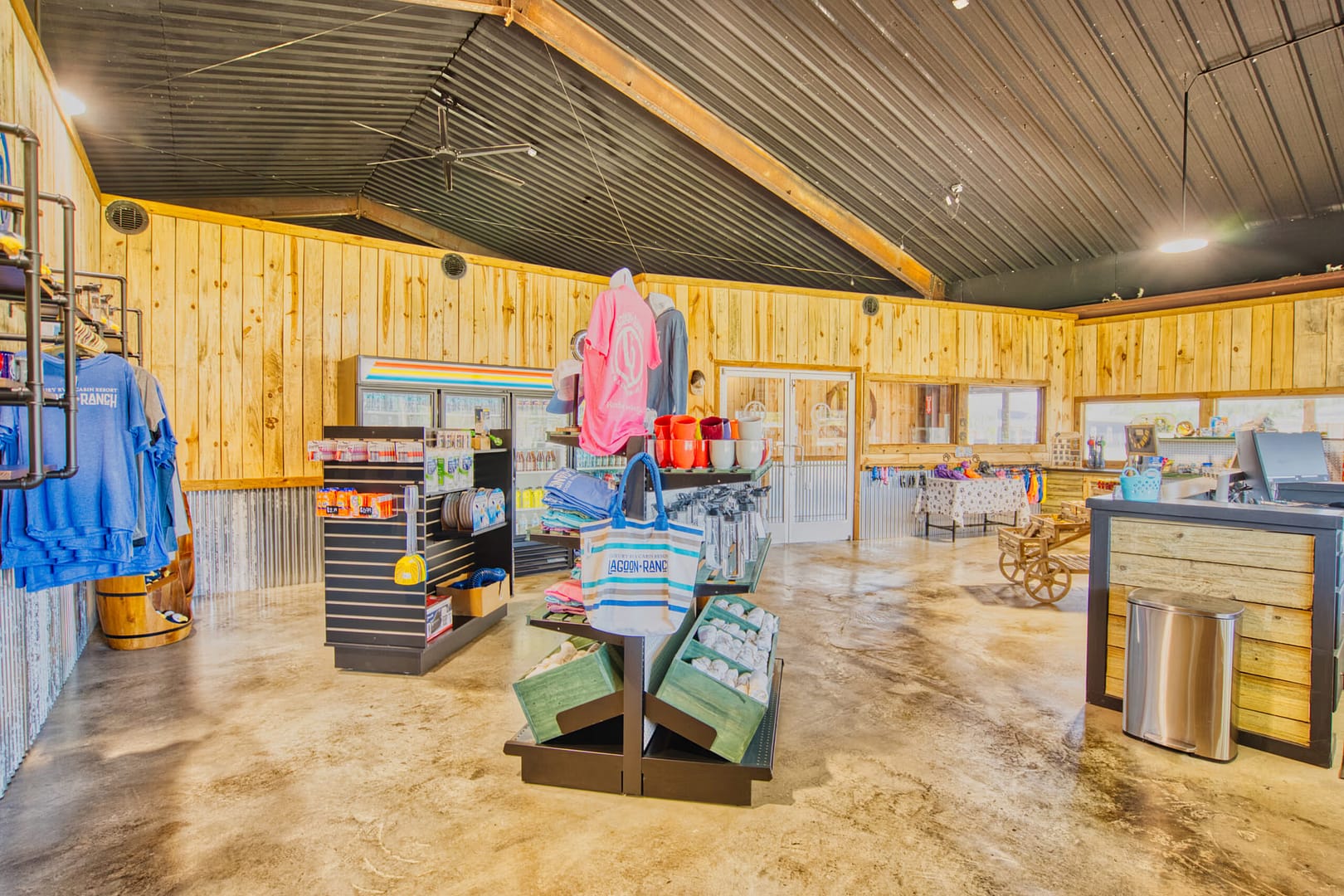 A rustic gift shop interior with wooden walls, merchandise on display racks, clothing, bags, and various items for sale, and a checkout counter on the right.