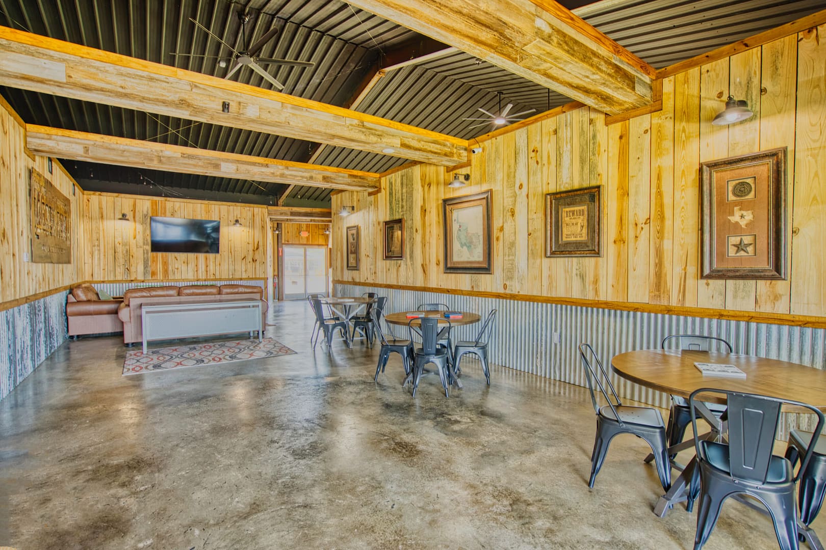 Rustic indoor seating area with wooden walls, metal chairs around round tables, framed art, a TV, and a sofa in the background on a polished concrete floor.