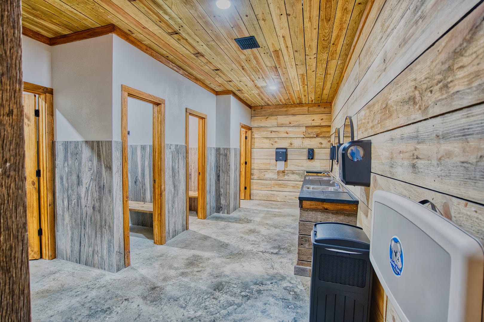 Modern public restroom with wooden walls and ceiling, stalls on the left, sinks and mirrors on the right, and a baby changing station in the foreground.