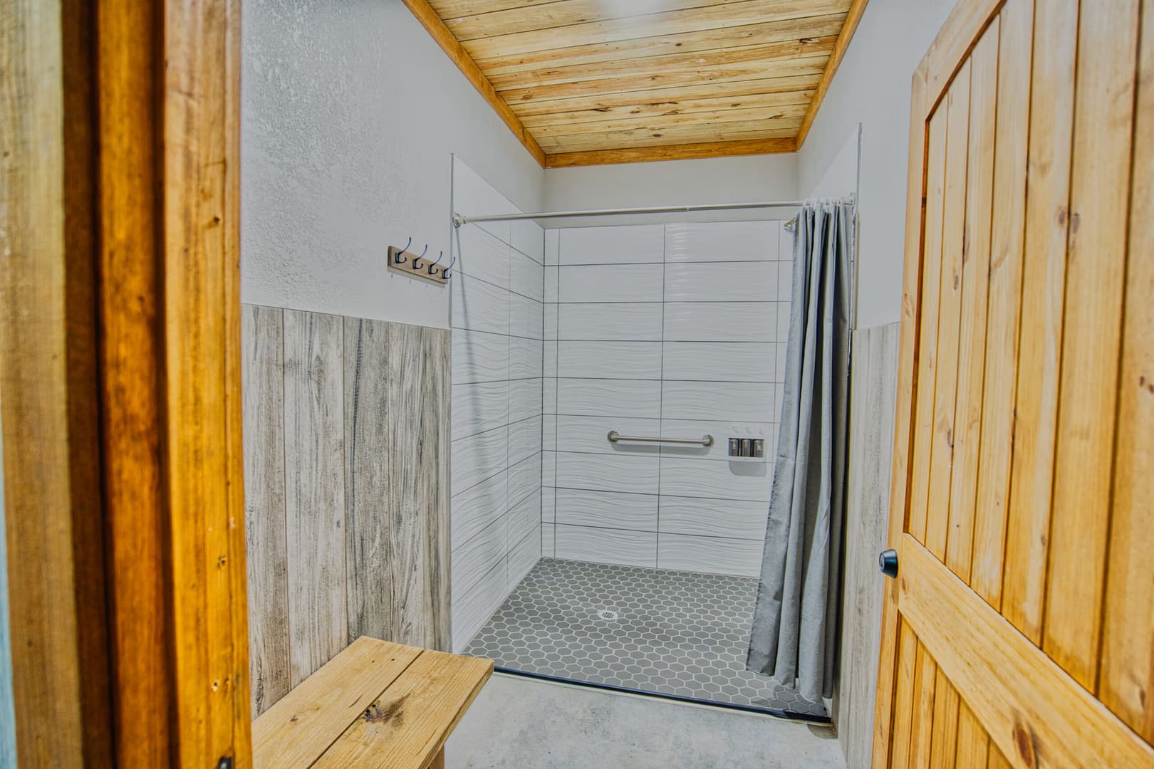 Accessible shower with grab bar, gray curtain, tiled walls, and wooden accents, including a door, bench, and ceiling. Four hooks are mounted on the left wall.