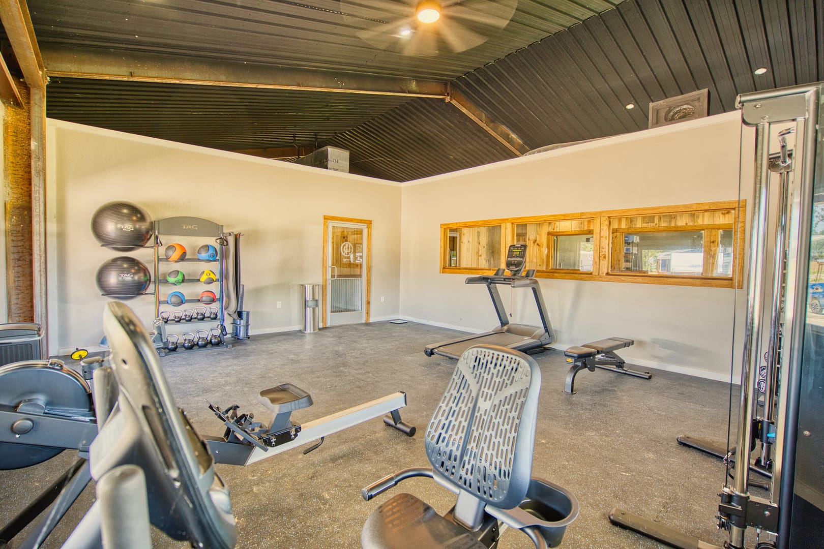 A gym room with cardio machines, weight rack, medicine balls, a bench, and large windows providing natural light.