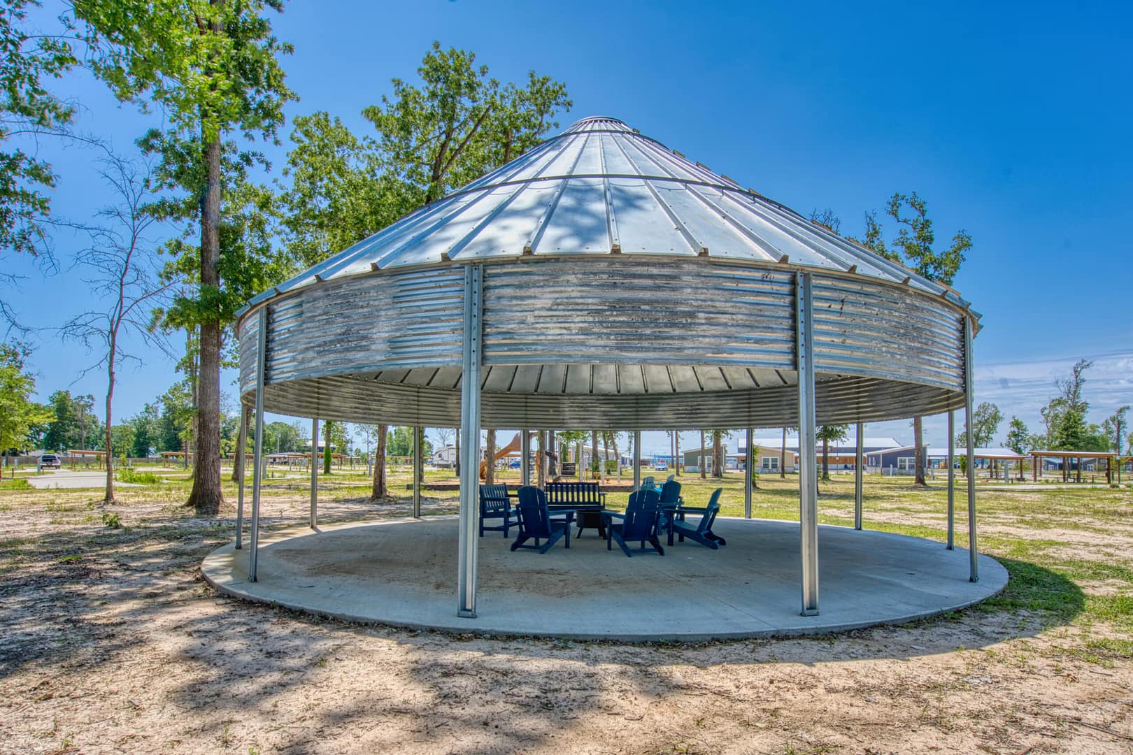 A round metal pavilion with open sides shelters a circle of blue chairs and a table, set on a concrete pad in a park with trees and clear sky.