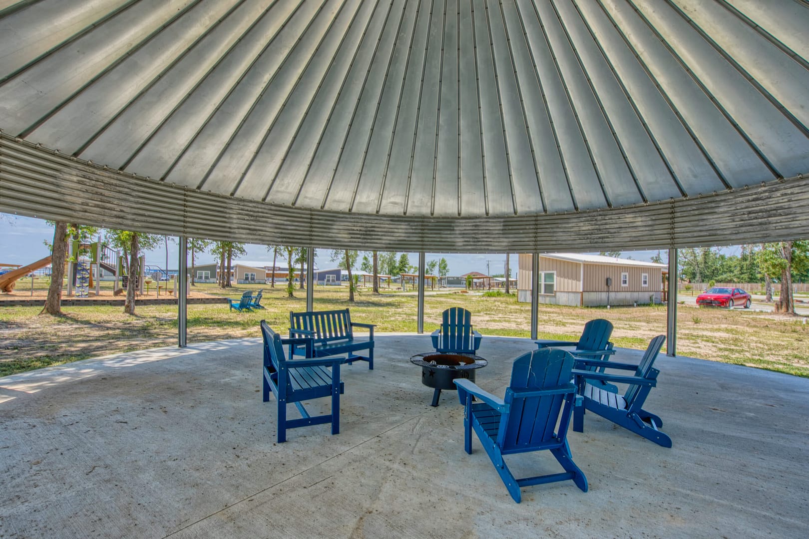 Six blue Adirondack chairs surround a fire pit under a large, open-sided metal pavilion, with buildings and trees visible outside.
