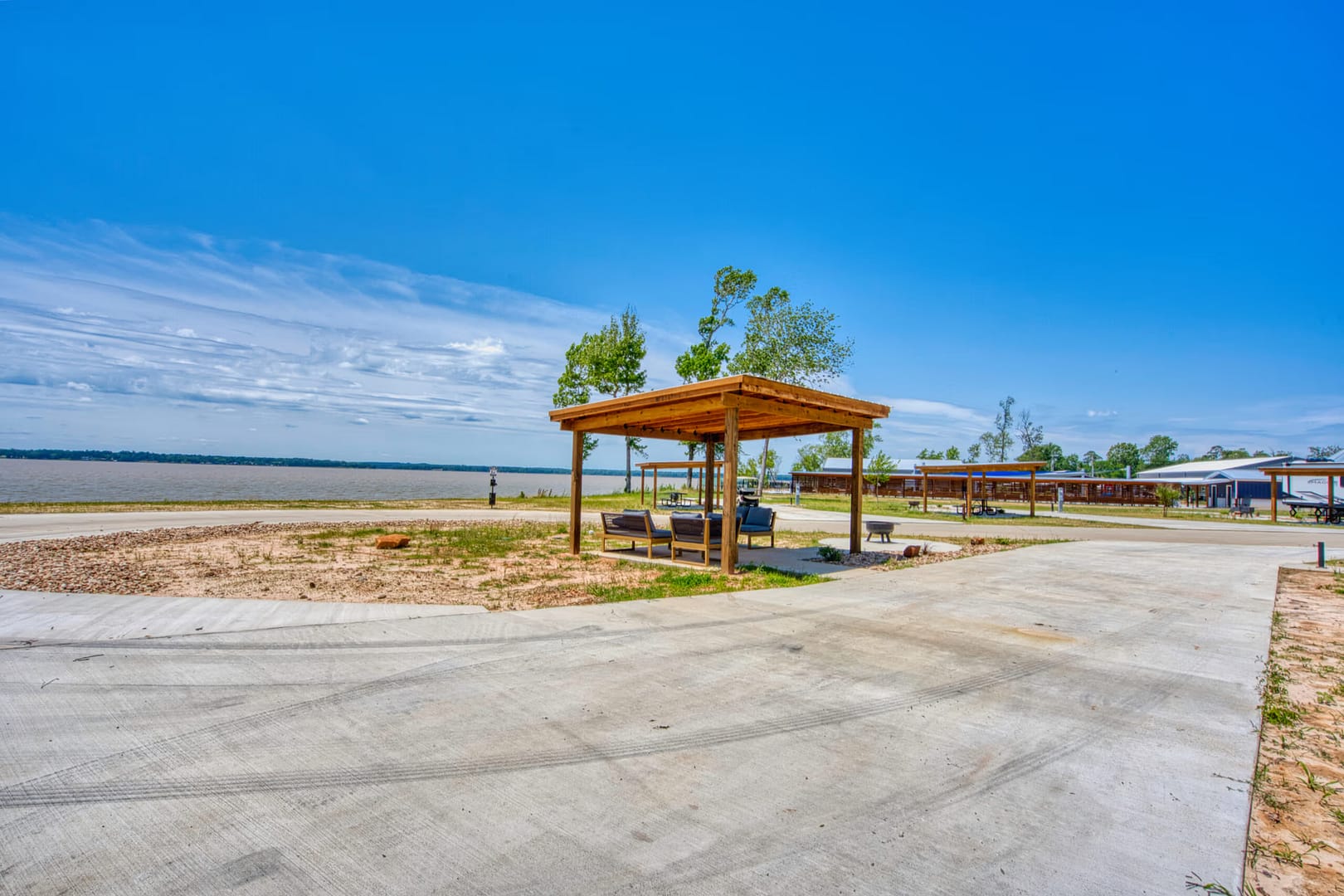 A wooden gazebo with outdoor seating is set on a concrete lot near a sandy, open landscape under a clear blue sky.