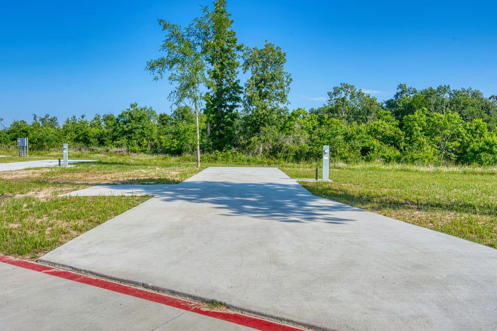 A concrete RV parking pad with utility hookups is surrounded by grass and trees under a clear blue sky.