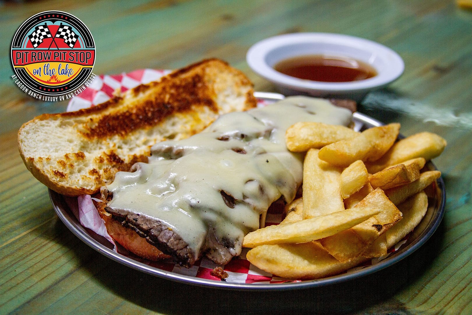A sandwich with melted cheese and beef, served with French fries and a side of dipping sauce on a plate; a "Pit Row Pit Stop" logo is visible in the image.