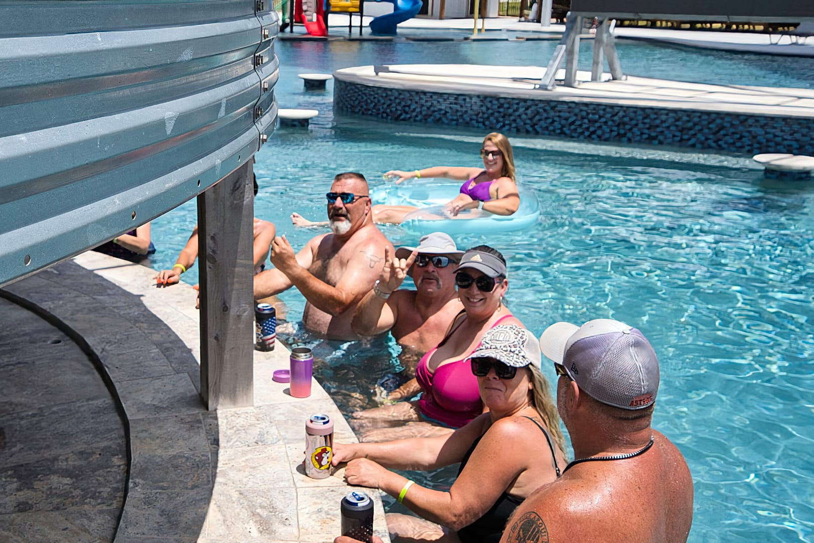 A group of adults sit and relax with drinks at the edge of a pool, while one person floats on an inflatable tube in the background.